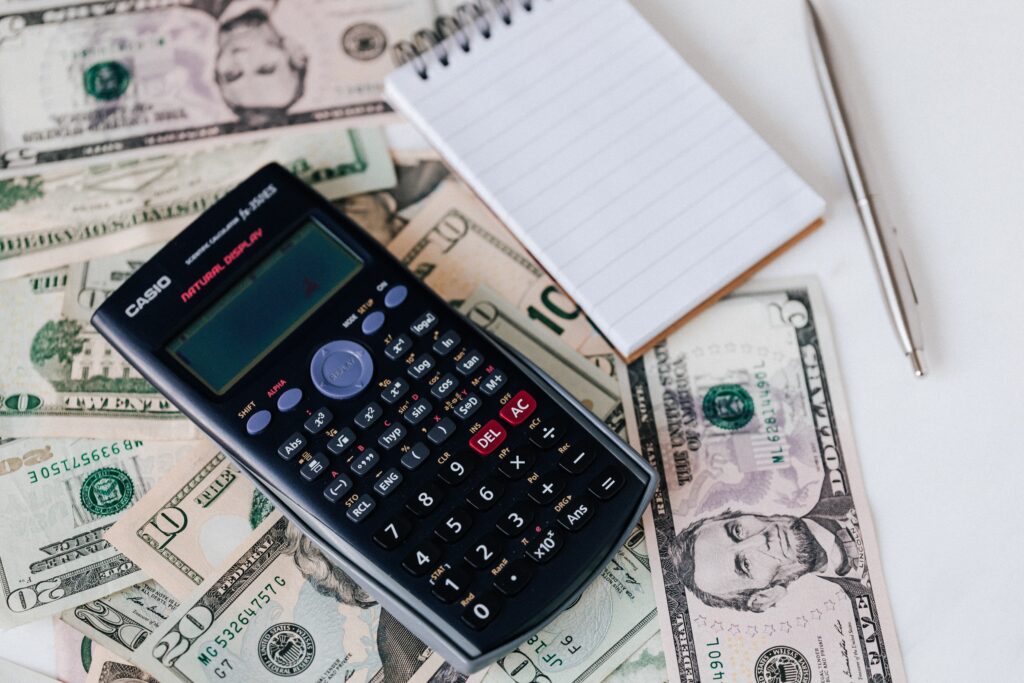 calculator on top of a pile of money beside a blank notepad and pen