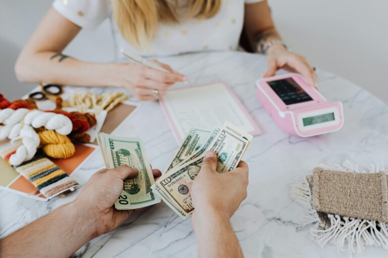 one hand holding cash, facing a person with a calculator and a spreadsheet. Two people doing budgeting activities.