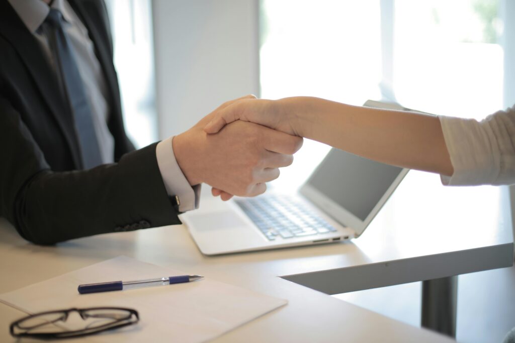 two hands shaking in front of an open laptop with a pen and glasses sitting on desk